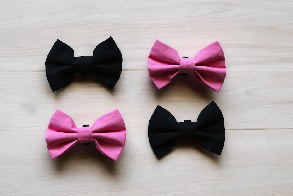 Four formal dog bowties in block colours - two pink and two black - arranged on a wooden tabletop.