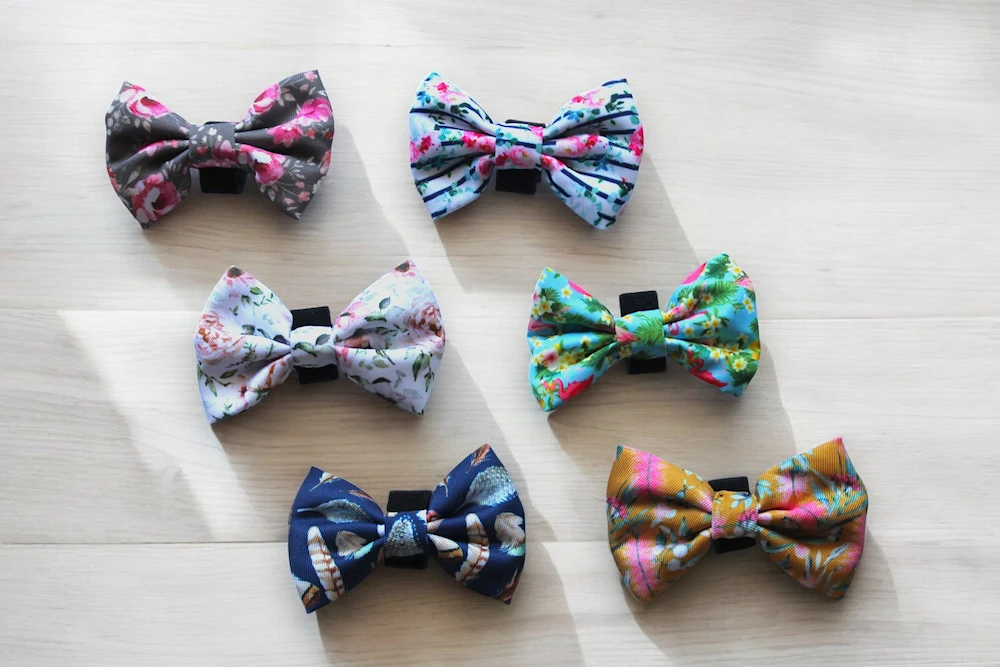 Six dog bowties of various multicolour patterns arranged on a wooden tabletop.
