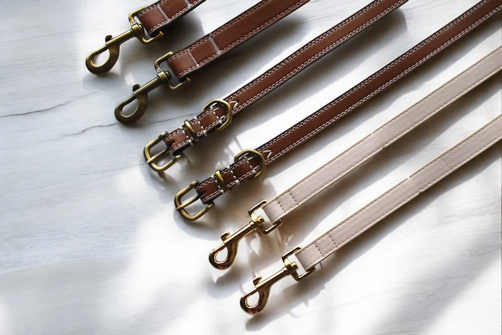 Six leather dog leads and collars with brass/gold highlights, lined up side by side on a wooden tabletop.